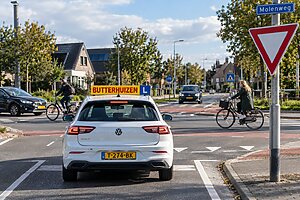Leerling beoordeelt tijdens rijles een drukke Nederlandse verkeerssituatie met kruispunt, fietsers, voetgangers en verkeersborden