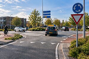 Leerling nadert tijdens rijles een Nederlandse rotonde met auto's, fietsers en voorrangsdriehoeken